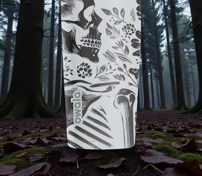 May include: A white Owala water bottle featuring a black skull and floral design. The bottle has a black and white skeletal illustration and the Owala logo. The background is a forest scene with tall trees and fallen leaves.