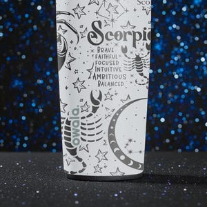 May include: A white Owala water bottle featuring a Scorpio zodiac design. The design includes a scorpion, stars, and the words "Brave, Faithful, Focused, Intuitive, Ambitious, Balanced." The bottle is set against a dark blue background with bokeh lights.