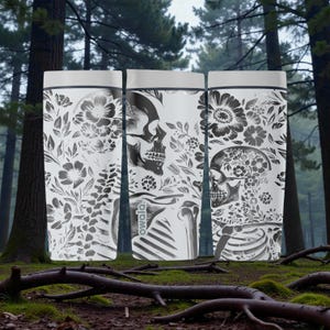 May include: Three white water bottles with a black and white floral and skull design. The bottles have a gray top and a black outline. The design includes flowers, leaves, and a human skull. The bottles are in a forest setting.