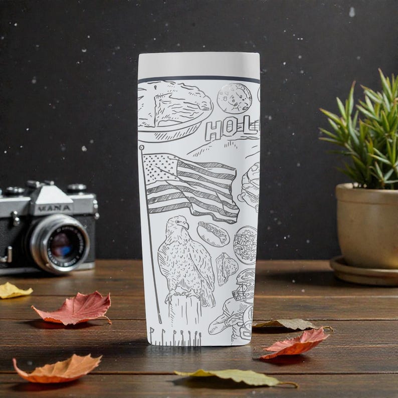 May include: White travel mug with a black and white illustration of American icons, including the American flag, an eagle, and a football player. The word "HOL" is visible on the mug. The mug is on a wooden surface.