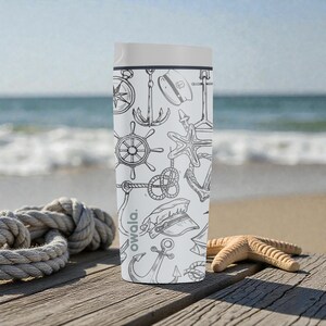 May include: A white Owala water bottle with a nautical-themed design. The bottle features black line drawings of anchors, compasses, ship wheels, and starfish. The bottle is on a wooden surface near the ocean.