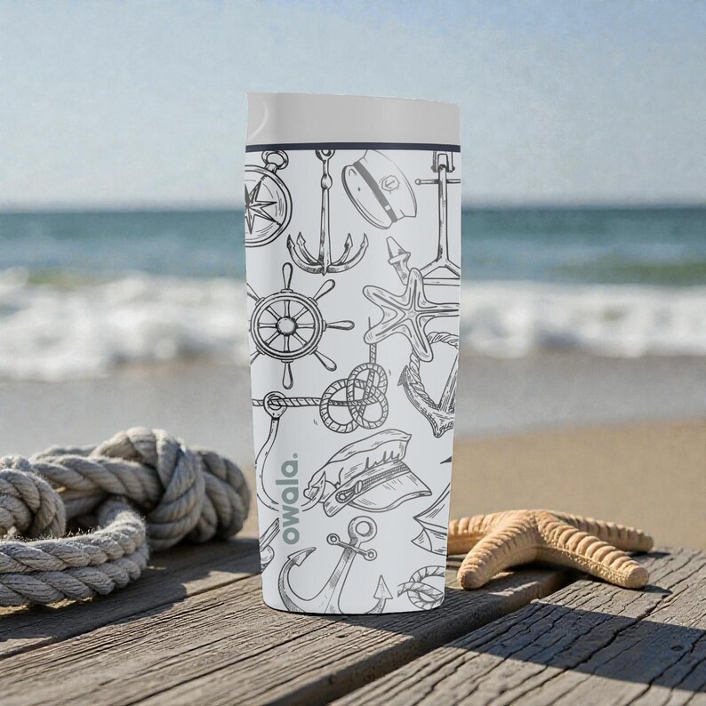May include: A white Owala water bottle with a nautical-themed design. The bottle features black line drawings of anchors, compasses, ship wheels, and starfish. The bottle is on a wooden surface near the ocean.