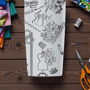 May include: A white, rectangular tumbler featuring a black line drawing of sewing tools and floral designs. The tumbler is surrounded by colorful fabric scraps, spools of thread, and orange scissors on a wooden surface.
