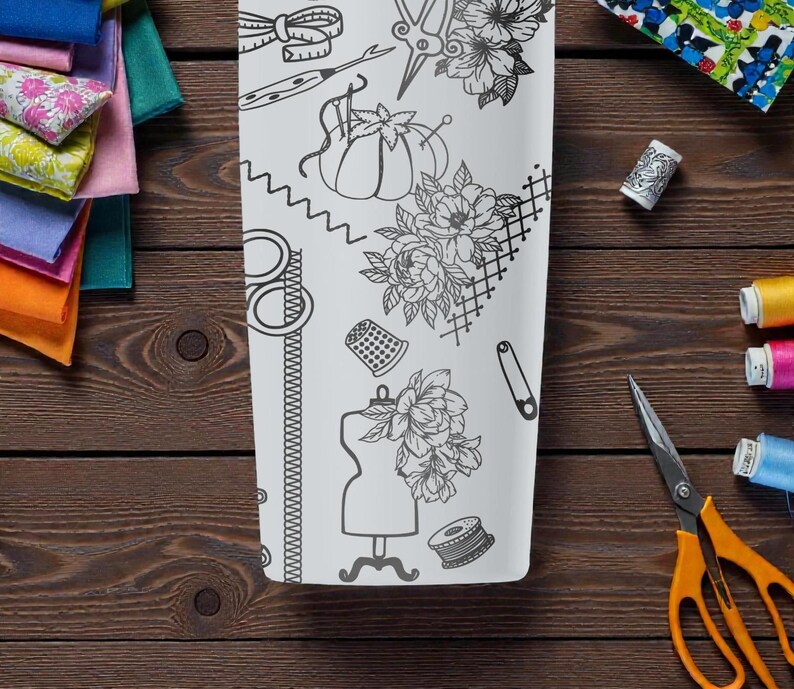 May include: A white, rectangular tumbler featuring a black line drawing of sewing tools and floral designs. The tumbler is surrounded by colorful fabric scraps, spools of thread, and orange scissors on a wooden surface.