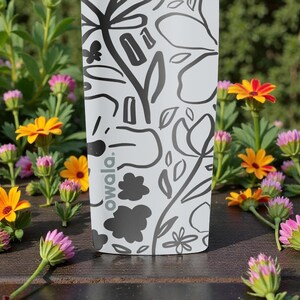 May include: A white water bottle with a black floral design and the word "owala." The bottle is surrounded by colorful flowers with yellow, orange, and pink petals. The bottle is on a wooden surface.