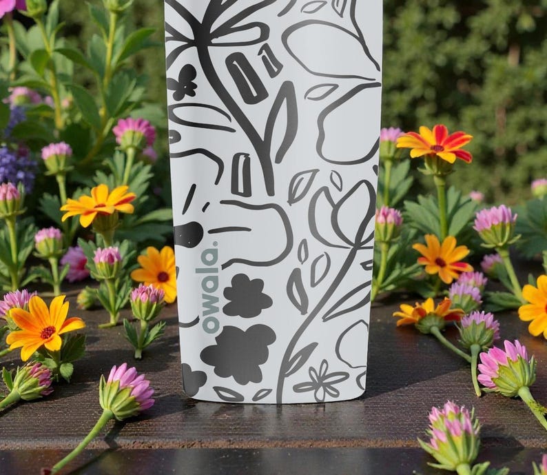 May include: A white water bottle with a black floral design and the word "owala." The bottle is surrounded by colorful flowers with yellow, orange, and pink petals. The bottle is on a wooden surface.
