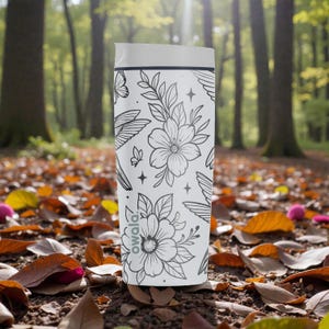 Puede incluir: Un vaso de viaje blanco con un dise&ntilde;o floral y de mariposas en tinta negra. El vaso tiene una tapa blanca y la marca "owala" est&aacute; impresa en el lateral. El fondo es un suelo forestal con hojas ca&iacute;das.