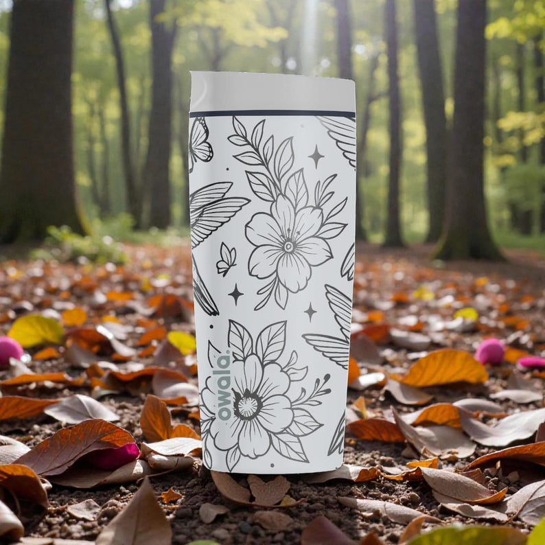 Puede incluir: Un vaso de viaje blanco con un dise&ntilde;o floral y de mariposas en tinta negra. El vaso tiene una tapa blanca y la marca "owala" est&aacute; impresa en el lateral. El fondo es un suelo forestal con hojas ca&iacute;das.