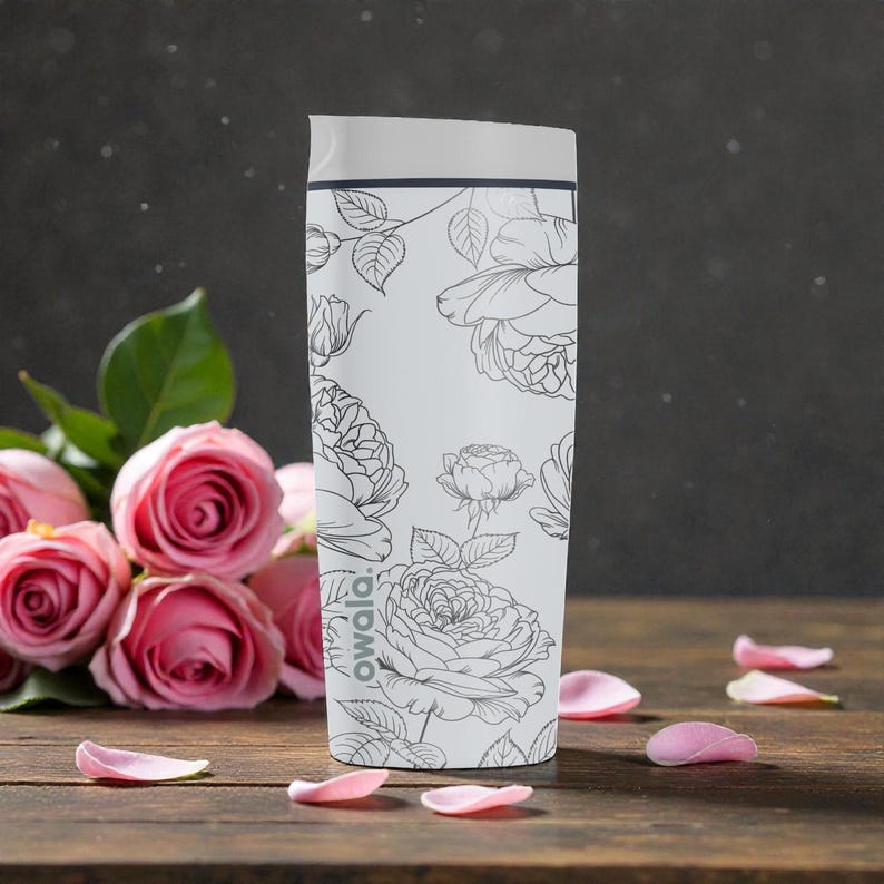 Op de afbeelding: Een witte Owala ge&iuml;soleerde waterfles met een bloemenpatroon. De fles heeft een zwart-witte lijntekening van rozen en bladeren. Roze rozenblaadjes liggen verspreid op een houten oppervlak naast een boeket roze rozen.