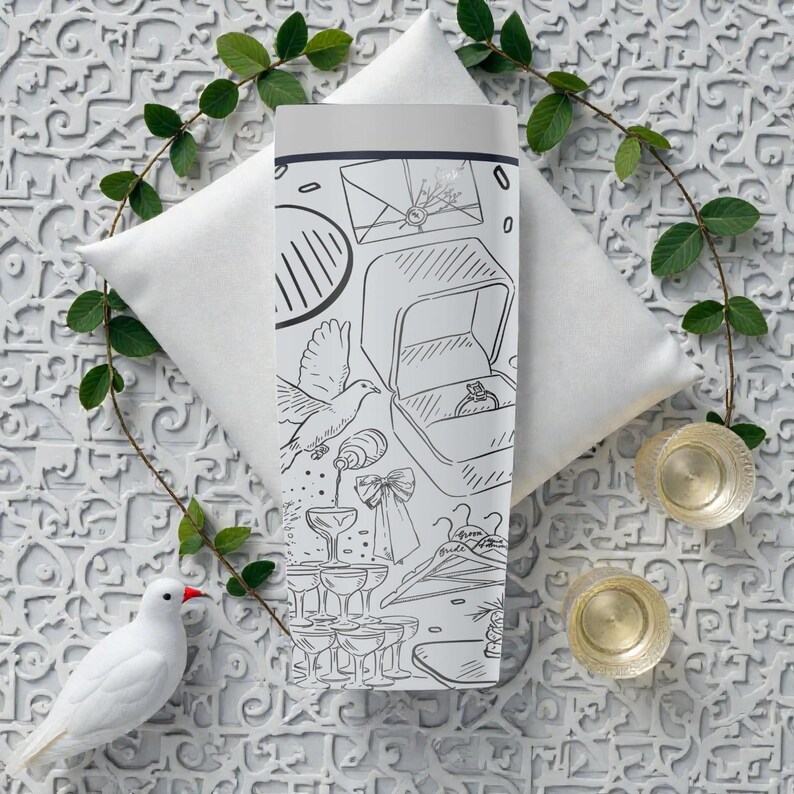 May include: A white tumbler with a black and white line drawing of wedding-related images, including a ring box, champagne glasses, and a dove. The tumbler is surrounded by a white pillow, greenery, and two gold-colored coasters.