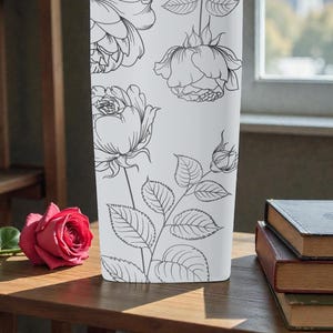 Op de afbeelding: Een witte rechthoekige vaas met zwarte lijntekeningen van rozen en bladeren. Een enkele rode roos ligt op een houten oppervlak. Boeken zijn op een tafel op de achtergrond gestapeld.