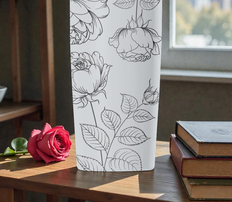 Op de afbeelding: Een witte rechthoekige vaas met zwarte lijntekeningen van rozen en bladeren. Een enkele rode roos ligt op een houten oppervlak. Boeken zijn op een tafel op de achtergrond gestapeld.
