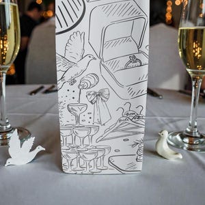 May include: A wedding reception table setting with two champagne flutes filled with a golden liquid. A decorative card with line art of wedding-related imagery, including a ring box, doves, and champagne glasses. Two small white dove figurines are on the table.