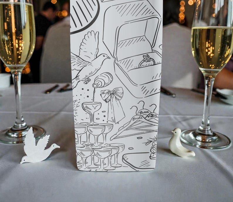 May include: A wedding reception table setting with two champagne flutes filled with a golden liquid. A decorative card with line art of wedding-related imagery, including a ring box, doves, and champagne glasses. Two small white dove figurines are on the table.