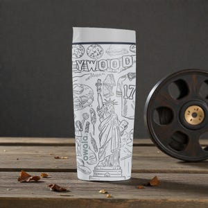 May include: White travel mug with a gray lid, featuring a black and white line drawing of Hollywood, the Statue of Liberty, and a football jersey with the number 17. The mug is on a wooden surface.