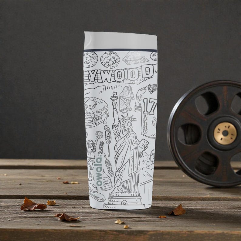 May include: White travel mug with a gray lid, featuring a black and white line drawing of Hollywood, the Statue of Liberty, and a football jersey with the number 17. The mug is on a wooden surface.