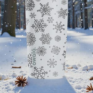Puede incluir: Una botella de agua blanca con un patr&oacute;n de copos de nieve negros. La botella tiene varios dise&ntilde;os de copos de nieve y la palabra "owala". La escena est&aacute; ambientada en un bosque nevado con pi&ntilde;as.