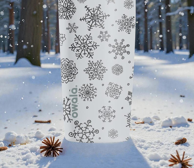 Puede incluir: Una botella de agua blanca con un patr&oacute;n de copos de nieve negros. La botella tiene varios dise&ntilde;os de copos de nieve y la palabra "owala". La escena est&aacute; ambientada en un bosque nevado con pi&ntilde;as.