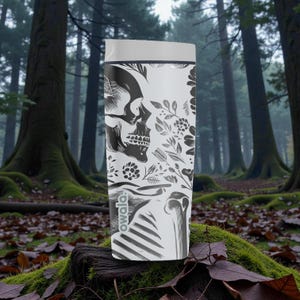 May include: A white Owala water bottle featuring a black and white skull design with floral accents. The bottle has a gray lid and is set against a forest backdrop. The Owala logo is visible on the lower portion of the bottle.