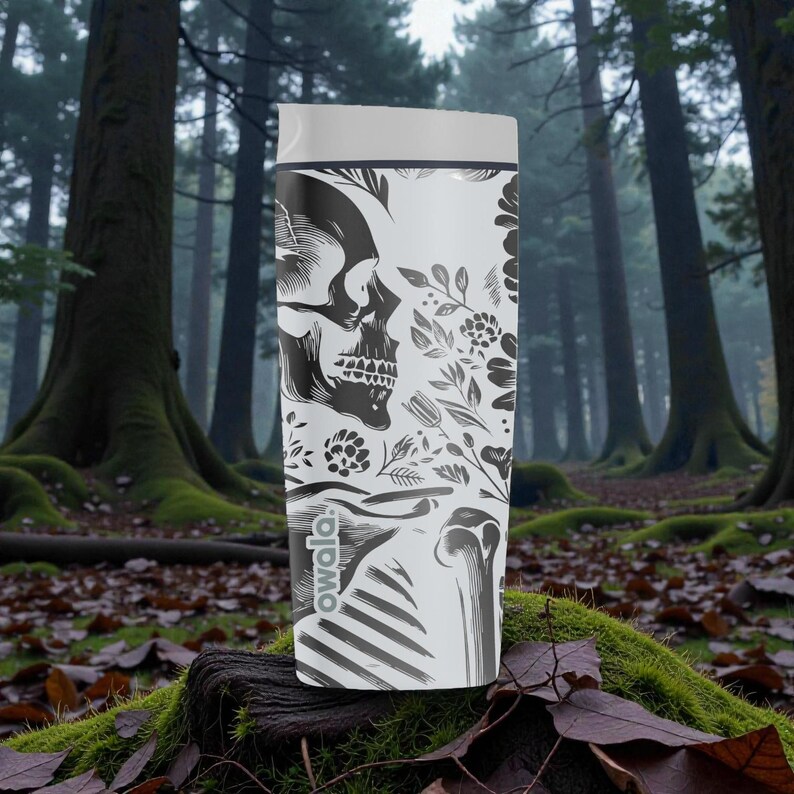 May include: A white Owala water bottle featuring a black and white skull design with floral accents. The bottle has a gray lid and is set against a forest backdrop. The Owala logo is visible on the lower portion of the bottle.