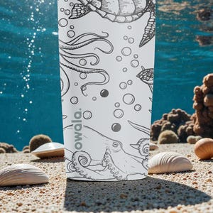 May include: A white water bottle with a black and white ocean-themed illustration featuring a turtle, octopus, and bubbles. The word "owala" is printed on the bottle. The bottle is on a sandy surface with seashells and coral in the background.