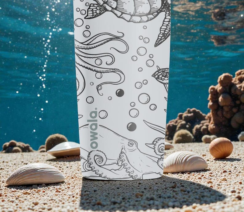 May include: A white water bottle with a black and white ocean-themed illustration featuring a turtle, octopus, and bubbles. The word "owala" is printed on the bottle. The bottle is on a sandy surface with seashells and coral in the background.