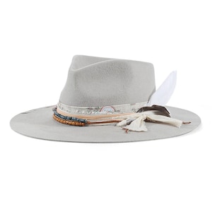 Könnte beinhalten: Ein hellgrauer Fedora-Hut mit breiter Krempe und eingedrückter Krone. Der Hut ist mit einem dekorativen Band verziert, das aus einem floralen Stoffstreifen, geschichteten Lederkordeln, blauen und braunen Perlen sowie zwei Federn besteht.
