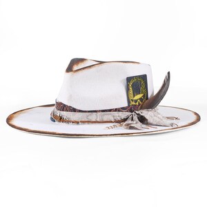 May include: A white fedora hat with a distressed brown scorched brim and crown. The hat features a decorative band made of layered fabric, a dark rectangular patch with a yellow skull graphic, and a single brown feather tucked into the side.