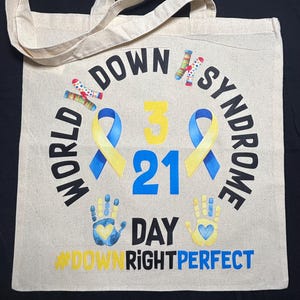 May include: Beige canvas tote bag with the words "WORLD DOWN SYNDROME DAY" in a circular design. The design includes blue and yellow ribbons, handprints, and the text "3 21". The hashtag "#DOWNRIGHTPERFECT" is also featured.