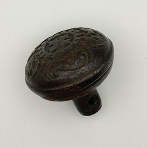 May include: A dark brown, antique-style door knob with an oval shape. The knob features an ornate, embossed floral design on its face and a small, cylindrical stem with a hole for mounting.