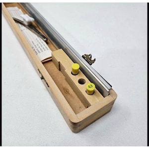 May include: A rectangular wooden storage box containing a long metal ruler, a metal scriber tool with a white handle, and two yellow cylindrical inserts, one of which is marked with the number 43, all nestled within custom-fitted interior slots.