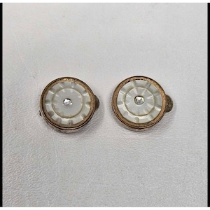 May include: A pair of vintage clip-on earrings. Each earring features a white, circular center with a small, clear rhinestone. The center is surrounded by a gold-toned metal frame with a decorative edge. The earrings are set against a white background.