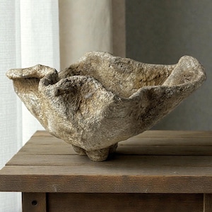 May include: A decorative, textured bowl with a natural, stone-like appearance. The bowl has an organic, irregular shape with a wide opening and sits on three small feet. The neutral color palette complements the rustic aesthetic.