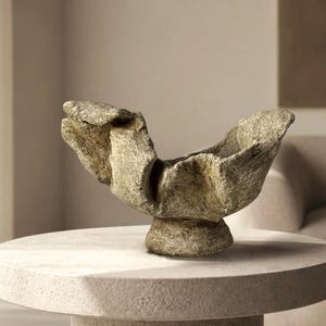Puede incluir: Un cuenco decorativo con una superficie rugosa y texturizada, con un acabado moteado en tonos beige y gris. El cuenco presenta una forma orgánica irregular con bordes anchos y acampanados y una base circular robusta, similar a la piedra.