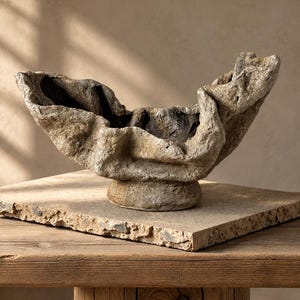 Peut inclure: Un bol décoratif avec une surface rugueuse et texturée rappelant la pierre dans des tons beige et marron. Le bol présente une forme organique irrégulière avec des plis profonds et une large ouverture, reposant sur une base circulaire.
