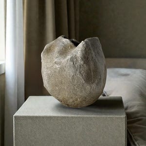May include: A sculptural ceramic vessel with a rough, textured surface in shades of beige and grey. The object features an irregular, organic shape with deep indentations and a wide, uneven opening at the top, showcasing a matte, earthy finish.