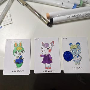 Puede incluir: Tres tarjetas de personajes dibujadas a mano con ilustraciones coloridas de personajes de animales. Las tarjetas est&aacute;n sobre una superficie blanca con materiales de arte en el fondo. Cada tarjeta tiene un nombre de personaje escrito en la parte inferior.