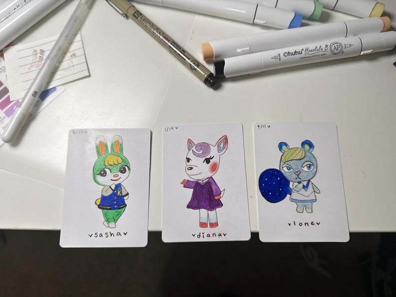 Puede incluir: Tres tarjetas de personajes dibujadas a mano con ilustraciones coloridas de personajes de animales. Las tarjetas est&aacute;n sobre una superficie blanca con materiales de arte en el fondo. Cada tarjeta tiene un nombre de personaje escrito en la parte inferior.