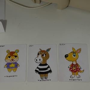 Puede incluir: Tres tarjetas de personajes ilustrados con Hamlet, Winnie y Carrie. Hamlet es un h&aacute;mster amarillo con una camisa morada con una estrella. Winnie es un caballo marr&oacute;n con una camisa a rayas blancas y negras. Carrie es un canguro amarillo con un vestido rojo.