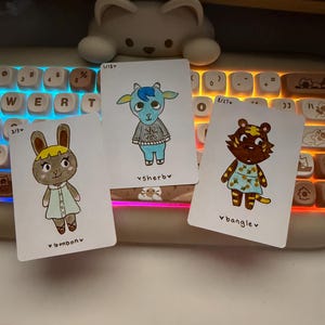 Puede incluir: Tres tarjetas de personajes ilustrados con un conejo, una cabra y un tigre, cada uno con atuendos y nombres &uacute;nicos. Las tarjetas est&aacute;n sobre un teclado con una luz blanca en forma de oso en el fondo. Las tarjetas tienen los nombres "bonbonv", "vsherby" y "banglev".