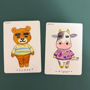 Puede incluir: Dos tarjetas ilustradas con personajes de dibujos animados. Una tarjeta muestra un oso pardo con una camisa a rayas amarillas y azules, con el texto "teddy" debajo. La otra tarjeta representa una vaca en blanco y negro con una camisa morada, con el texto "tipperv" debajo.