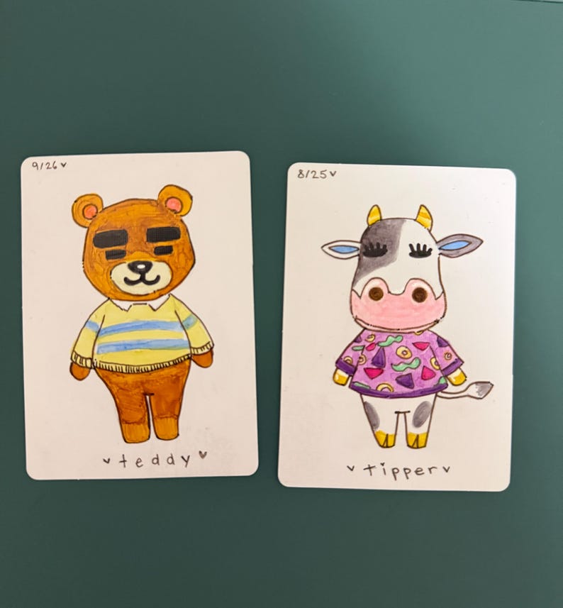 Puede incluir: Dos tarjetas ilustradas con personajes de dibujos animados. Una tarjeta muestra un oso pardo con una camisa a rayas amarillas y azules, con el texto "teddy" debajo. La otra tarjeta representa una vaca en blanco y negro con una camisa morada, con el texto "tipperv" debajo.