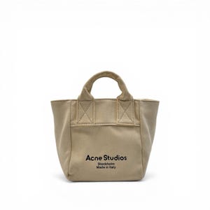 May include: A beige canvas tote bag with a small handle. The bag features the text "Acne Studios Stockholm Made in Italy" in black. The bag has a rectangular shape with reinforced stitching details.