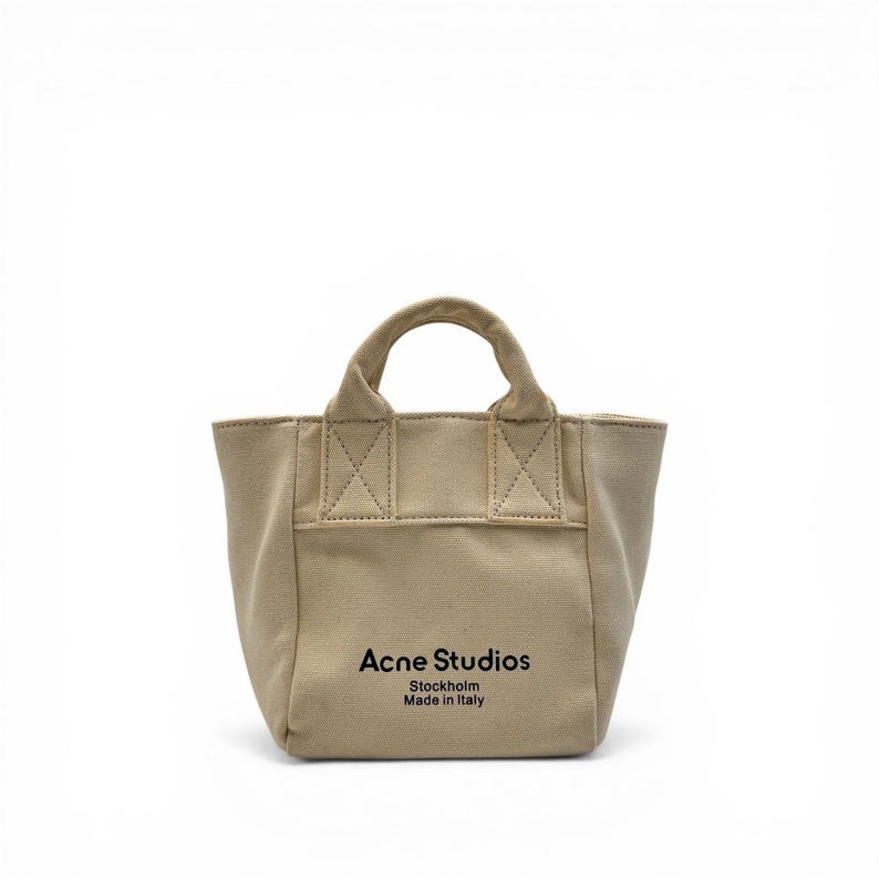 May include: A beige canvas tote bag with a small handle. The bag features the text "Acne Studios Stockholm Made in Italy" in black. The bag has a rectangular shape with reinforced stitching details.