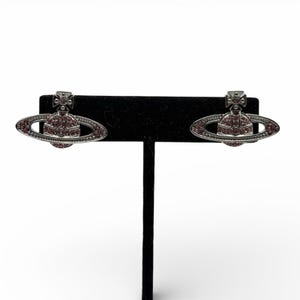 Pode incluir: Um par de brincos prateados com um design de planeta e uma cruz no topo. O planeta é circundado por um anel e preenchido com pequenas pedras preciosas rosa. Os brincos são exibidos em um suporte preto sobre um fundo branco.