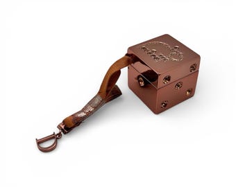May include: A copper-colored cube-shaped accessory with a brown strap and a metal 'D' charm. The cube has a decorative design on top and small circular accents on the sides. The strap is a mix of brown and silver.