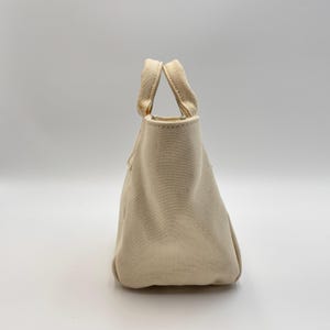 May include: A small, off-white canvas tote bag with two matching handles. The bag has a rectangular base and a slightly tapered shape, with a simple, minimalist design. The handles are securely attached to the top of the bag.