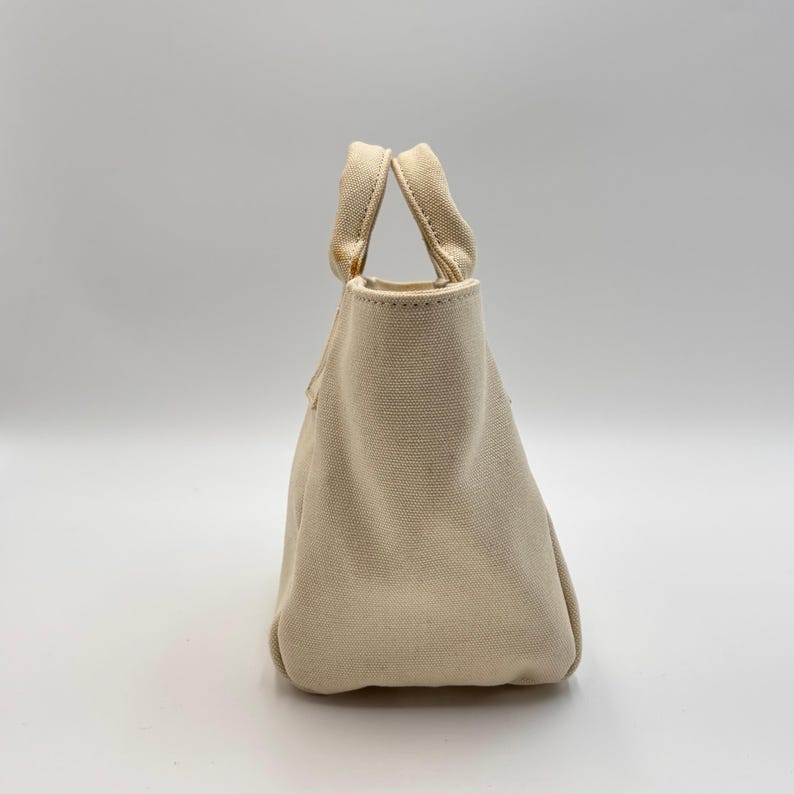 May include: A small, off-white canvas tote bag with two matching handles. The bag has a rectangular base and a slightly tapered shape, with a simple, minimalist design. The handles are securely attached to the top of the bag.