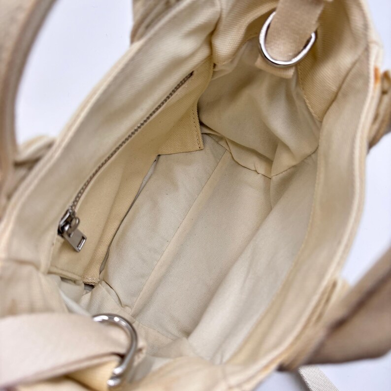 May include: Cream-colored canvas tote bag with a zippered interior pocket and silver-toned hardware. The bag has a shoulder strap and a top handle. The interior is a lighter shade of cream.