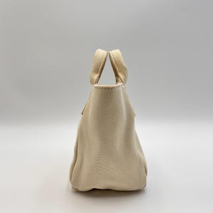 May include: A small, beige canvas tote bag with two sturdy handles. The bag has a simple, rounded shape and a textured surface. The handles are attached at the top of the bag, and the bag appears to be empty.